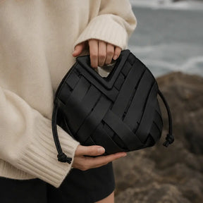 Detalhe em close da bolsa Iris preta da Nualle sendo segurada pela alça triangular geométrica, destacando o acabamento premium do couro trançado, com a modelo vestindo um suéter off-white em um cenário costeiro