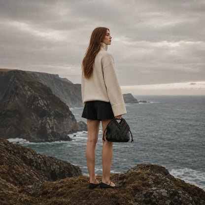 Uma mulher jovem, de costas e com cabelo ruivo comprido, está em pé na borda de um penhasco costeiro íngreme, olhando para o mar agitado sob um céu nublado. Ela veste um suéter de gola alta creme oversized e shorts pretos. Na mão esquerda, ela segura a bolsa Nualle 'Iris' de couro trançado preto, com sua alça estruturada e cordões laterais