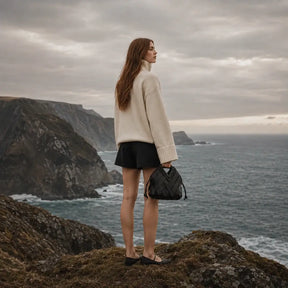 Uma mulher jovem, de costas e com cabelo ruivo comprido, está em pé na borda de um penhasco costeiro íngreme, olhando para o mar agitado sob um céu nublado. Ela veste um suéter de gola alta creme oversized e shorts pretos. Na mão esquerda, ela segura a bolsa Nualle 'Iris' de couro trançado preto, com sua alça estruturada e cordões laterais