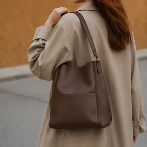 Modelo feminina segurando a Bolsa de Ombro em Couro Eva Coffee em ambiente lifestyle com luz natural e tons terrosos