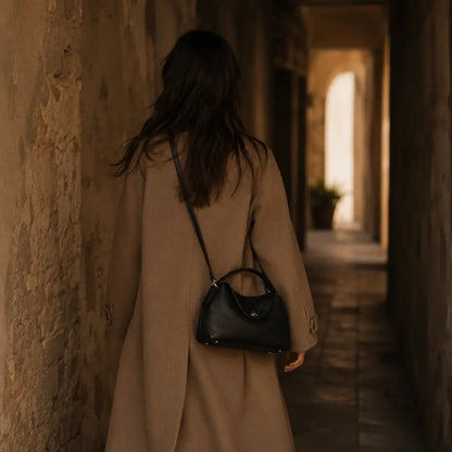 Bolsa Feminina em Couro Calla Preta usada no ombro em modo crossbody por modelo com trench coat camel em corredor de pedra