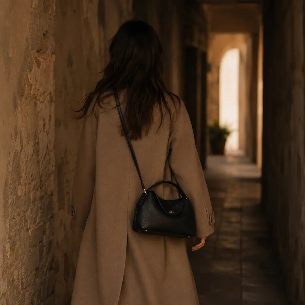 Bolsa Feminina em Couro Calla Preta usada no ombro em modo crossbody por modelo com trench coat camel em corredor de pedra
