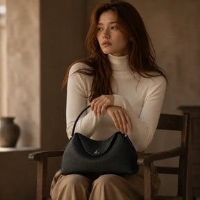 Modelo feminina sentada em cadeira de madeira com gola alta creme segurando a Bolsa Feminina em Couro Calla Preta no colo