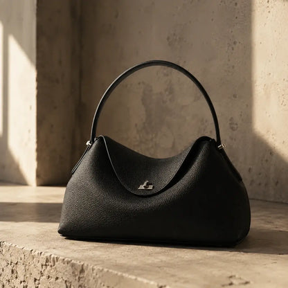 Bolsa Feminina em Couro Calla Preta com alça arqueada slim e fecho prata apoiada em superfície de concreto com luz natural