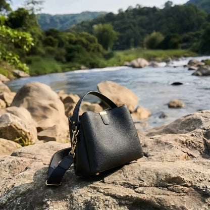 Bolsa feminina em couro preta com alça transversal e acabamento premium, estilo luxo moderno em cenário natural com pedras e lago