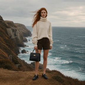 Modelo segurando a Bolsa Feminina em Couro Preta Louise pela alça de mão, combinada com suéter creme e shorts marrom em cenário costeiro