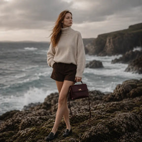 Modelo observando o mar com a Bolsa Feminina em Couro Louise Bordô em cenário costeiro cinematográfico.