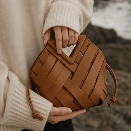 Bolsa feminina marrom caramelo de mão em couro trançado estilo intrecciato, com foco na alça triangular rígida e recortada, segurada por uma mão em um cenário costeiro."