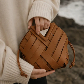 Bolsa feminina marrom caramelo de mão em couro trançado estilo intrecciato, com foco na alça triangular rígida e recortada, segurada por uma mão em um cenário costeiro."