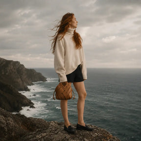 "Uma modelo de costas em pé num penhasco costeiro nublado, usando um suéter creme oversized e segurando uma bolsa feminina marrom caramelo em couro trançado.