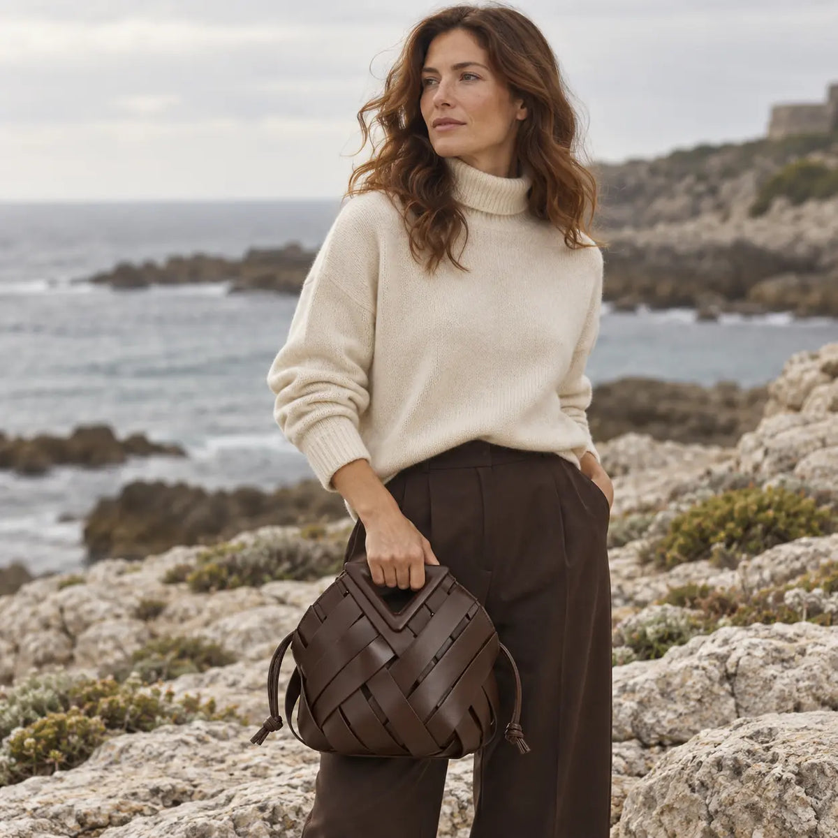 Mulher elegante com a Bolsa Iris Coffee em cenário editorial na costa rochosa