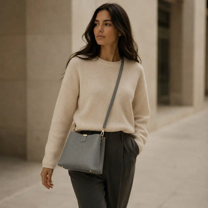 Modelo feminina caminhando em ambiente urbano com suéter bege usando a Bolsa de Mão em Couro Elara Cinza no modo crossbody com alça transversal