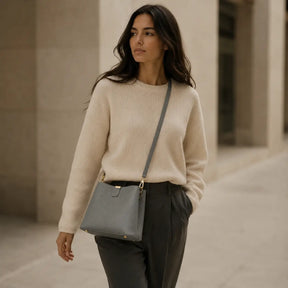 Modelo feminina caminhando em ambiente urbano com suéter bege usando a Bolsa de Mão em Couro Elara Cinza no modo crossbody com alça transversal