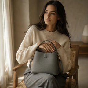 Modelo feminina sentada em cadeira de madeira com suéter creme segurando a Bolsa de Mão em Couro Elara Cinza no colo com alça de mão