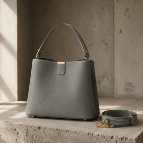 Bolsa de Mão em Couro Elara Cinza apoiada em superfície de concreto com alça transversal ao lado, ferragens douradas e luz natural lateral