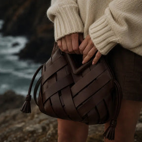 Bolsa de mão em couro trançado marrom escuro com alça triangular e pingentes tassel, segurada por mulher com suéter creme sobre fundo de costa rochosa
