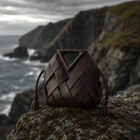 Bolsa de mão trançada em couro marrom escuro com alça triangular e cordões tassel apoiada sobre rocha com penhascos e oceano ao fundo