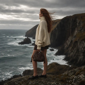 Mulher ruiva de costas com suéter oversized creme e shorts marrom, segurando bolsa trançada de couro marrom em penhasco com mar agitado ao fundo

